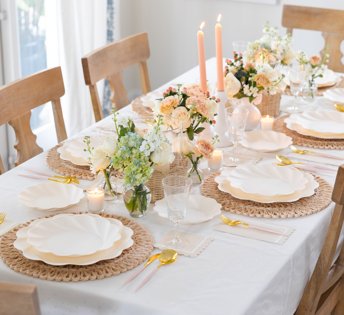 A dining table set with Sophistiplate LLC's Eco Cream & Blush Cutlery Bundle, woven placemats, bamboo plates, glassware, floral centerpieces, and candles creates an elegant and inviting atmosphere.