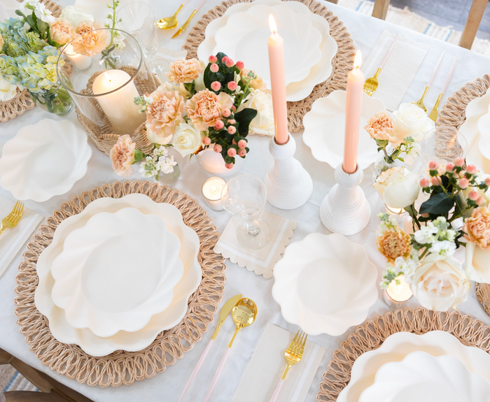 Elegant table setting with the Eco Cream & Blush Cutlery Bundle by Sophistiplate LLC, paired with white scalloped plates, pink candles, floral centerpieces of roses and carnations, woven placemats, glassware, and a white tablecloth.
