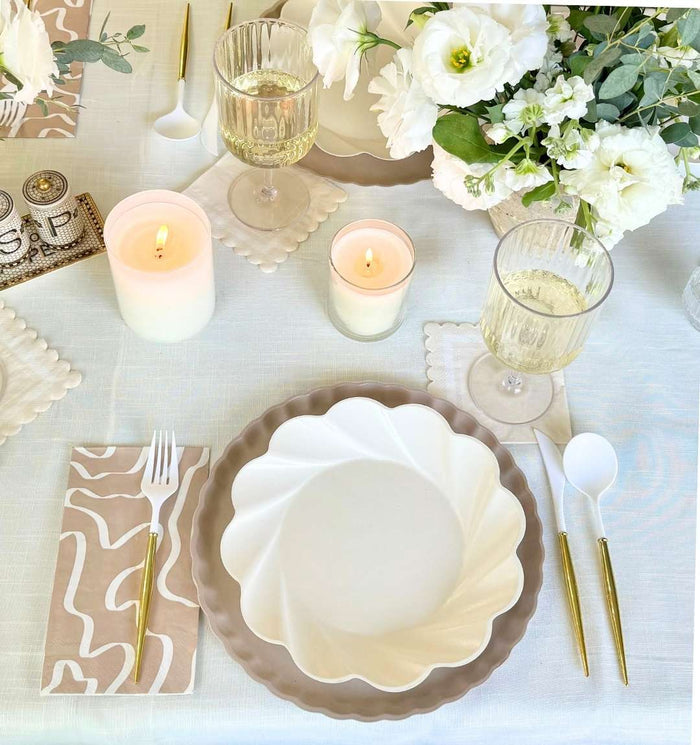 Create an inviting tablescape with Sophistiplate's Eco-Friendly Table Setting in Cream, featuring gold cutlery, patterned napkins, wine glasses, candles, and a white floral centerpiece on a cream tablecloth.