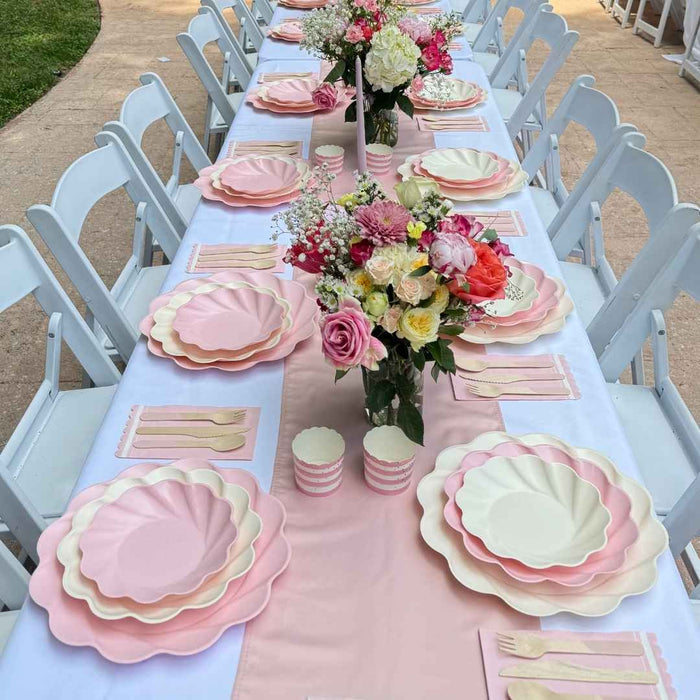 A long outdoor table is styled with Sophistiplate LLC's Eco Cream & Blush Table Setting for 16, including white chairs, party plates, wooden cutlery, striped cups, and floral centerpieces on a pink and white runner.