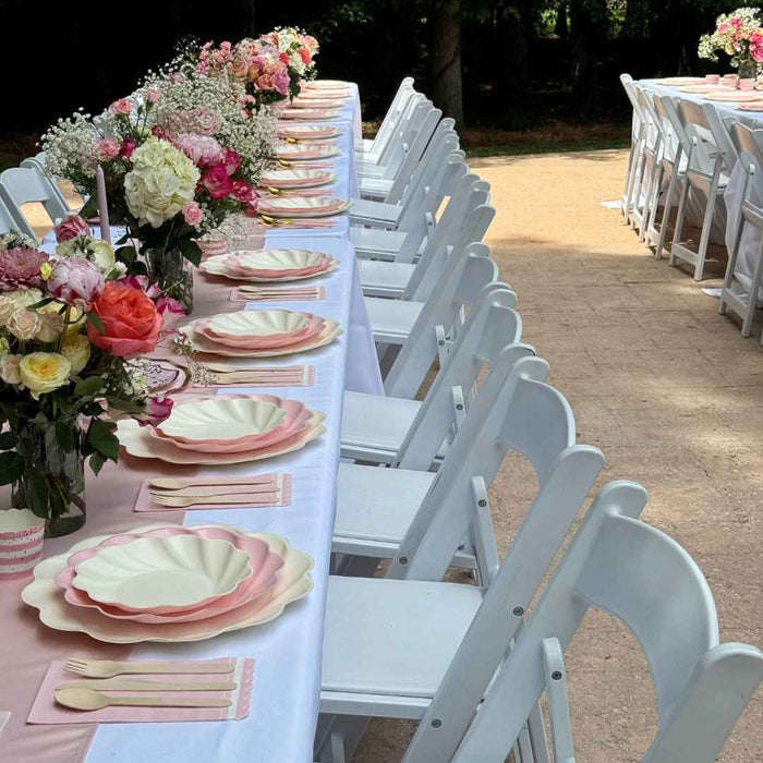 An outdoor table features the Eco Cream & Blush Table Setting for 16 by Sophistiplate LLC, elegantly arranged with disposable plates and wooden cutlery, while white folding chairs line both sides.