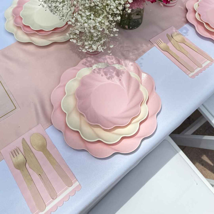 Create a pastel tablescape with the Sophistiplate LLC Eco Cream & Blush Table Setting for 16—scalloped pink and cream plates, pink napkins with gold cutlery prints, a white tablecloth, and white baby’s breath centerpiece.