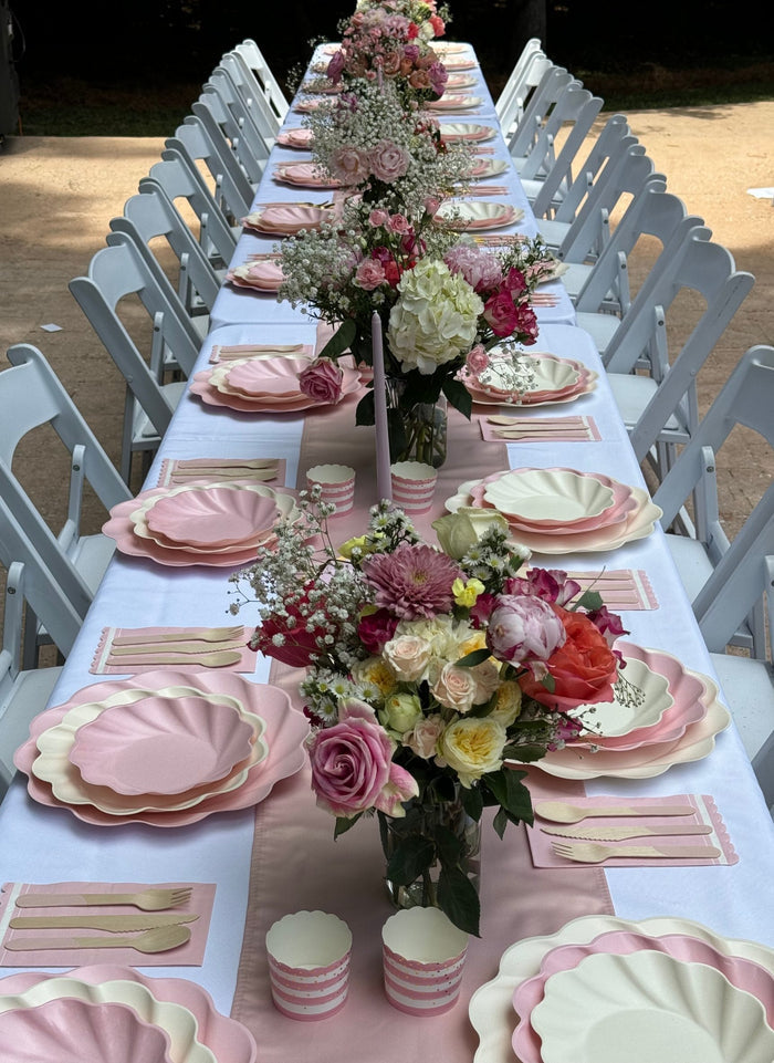 An outdoor table features the Eco-Friendly Table Setting- Blush by Sophistiplate LLC: white and pink scalloped compostable plates, gold cutlery, pink-striped cups, floral rose centerpieces, and white chairs on both sides.