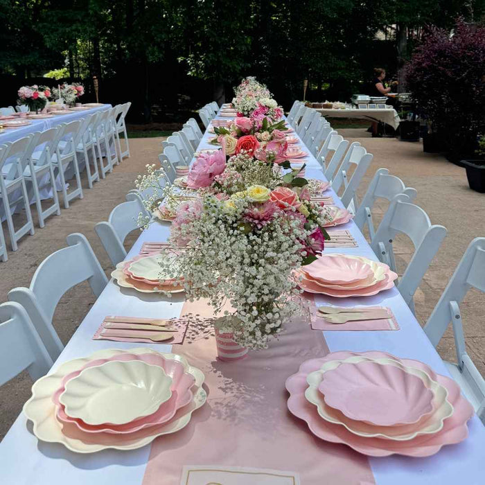 A long outdoor table is set with Sophistiplate LLC's Eco Cream & Blush Table Setting for 16, featuring white and pink eco-friendly plates, matching napkins, floral centerpieces, and surrounded by white chairs amidst greenery.