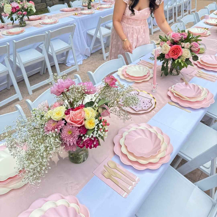 A woman in a pink dress arranges flowers at an outdoor table set with Sophistiplate LLC’s Eco Cream & Blush Table Setting for 16, featuring pastel eco-friendly plates, gold cutlery, and floral centerpieces with roses and baby’s breath.