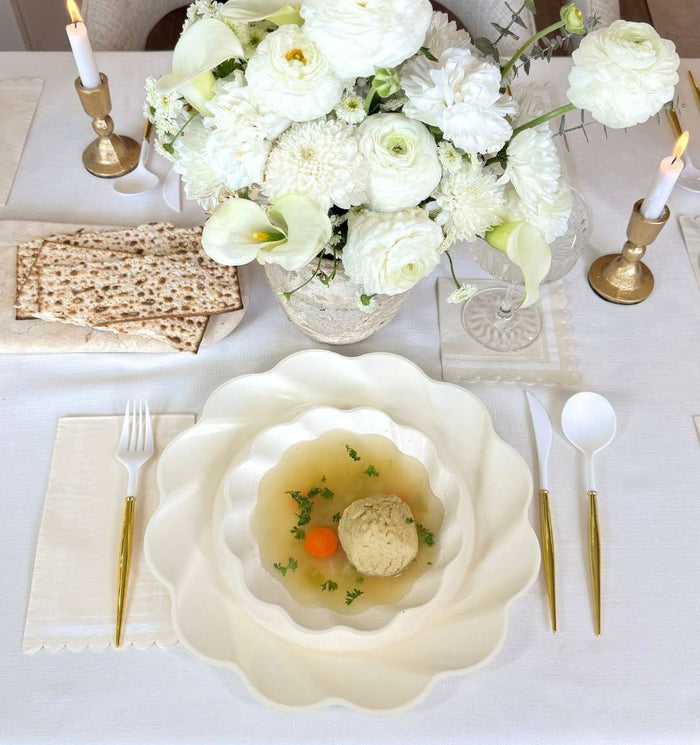 A white table featuring the Sophistiplate Eco-Friendly Table Setting- Cream: gold cutlery, stacked bamboo plates, matzo ball soup with carrots and herbs, matzo crackers, candles, and a white floral centerpiece.