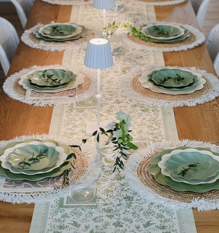The Eco Cream & Sage Table Setting by Sophistiplate LLC features layered green and cream eco-friendly plates on rustic placemats, a white floral table runner, small white lamps, and greenery accents for a stylish dining experience.