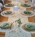 The Eco Cream & Sage Table Setting by Sophistiplate LLC features layered green and cream eco-friendly plates on rustic placemats, a white floral table runner, small white lamps, and greenery accents for a stylish dining experience.