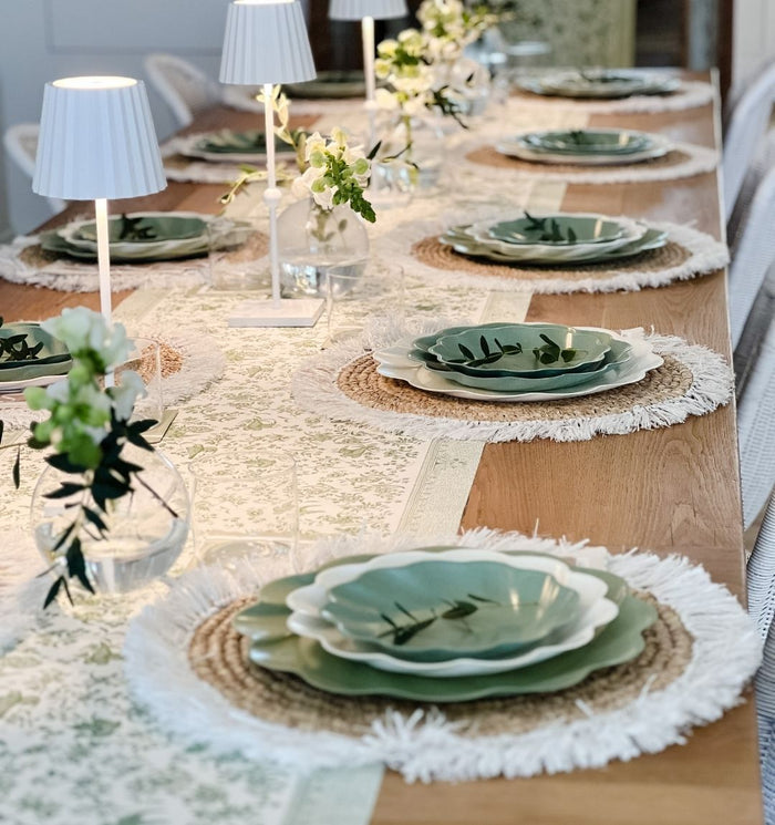 The Eco Cream & Sage Table Setting by Sophistiplate LLC features eco-friendly leaf-shaped plates, woven placemats, white fringed chargers, small vases with greenery, and white table lamps for an elegant tablescape.