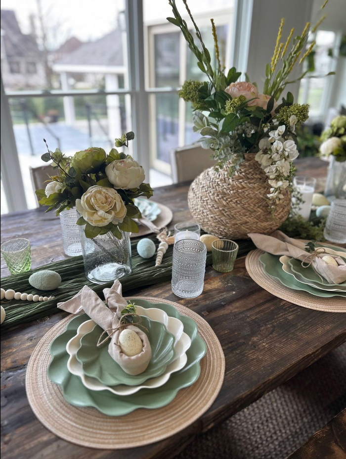 The Eco Cream & Sage Table Setting by Sophistiplate LLC features green and white eco-friendly plates, beige napkins, glassware, egg-shaped accents, and floral centerpieces in woven vases on a rustic table with large windows in the background.