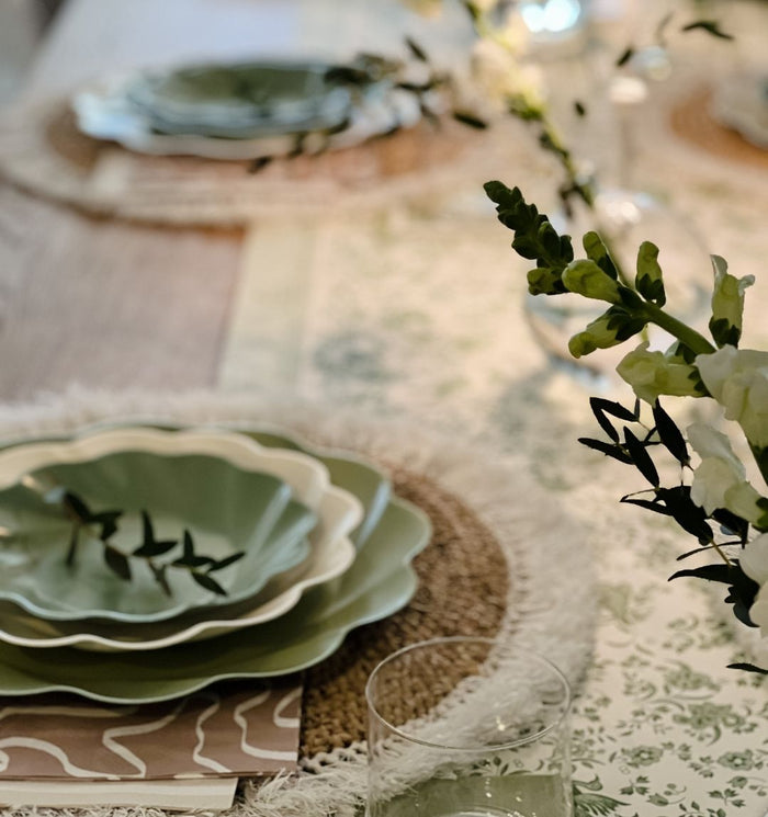 The Eco Cream & Sage Table Setting by Sophistiplate LLC features layered green and white eco-friendly plates on woven placemats, a floral runner, glassware, and fresh flowers for an elegant dining experience.