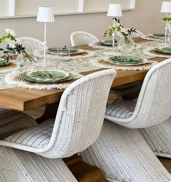 The Eco Cream & Sage Table Setting by Sophistiplate LLC features leaf-shaped plates, woven placemats, white wicker chairs, table lamps, and vases with white flowers for an elegant, fresh, and natural look.