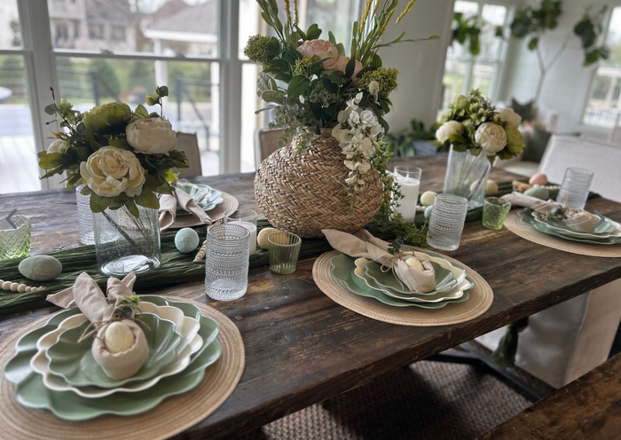 The Eco Cream & Sage Table Setting by Sophistiplate LLC features layered green and white eco-friendly plates, cloth napkins, glassware, and a woven vase with pink and white flowers. Decorative eggs and greenery complete this spring centerpiece.