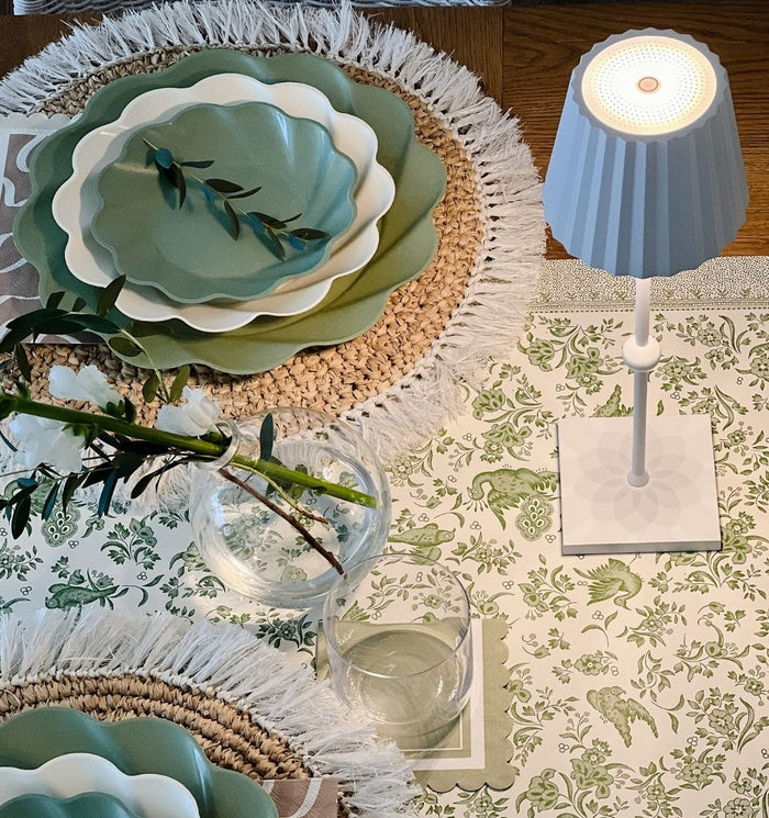 The Eco Cream & Sage Table Setting by Sophistiplate LLC features eco-friendly green and white plates, woven placemats, a water glass, a vase with greenery, and a white pleated-shade lamp on a floral tablecloth.