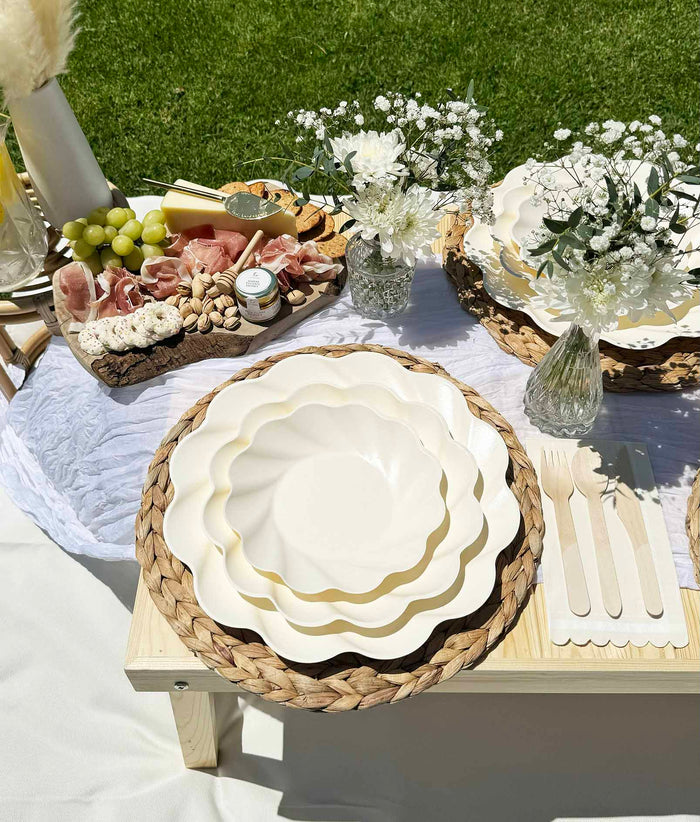 The Eco-Friendly Table Setting- Cream by Sophistiplate stylishly decorates a grassy picnic with compostable party plates, woven placemats, wooden cutlery, glass vases of white flowers, and a charcuterie board of grapes, meat, cheese, and nuts.