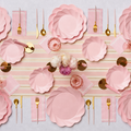 A table set with the Eco-Friendly Table Setting- Blush by Sophistiplate LLC, featuring compostable party plates with pink scalloped edges, gold flatware, pink napkins, pink glasses, and a centerpiece of white and pink flowers atop a striped table runner.