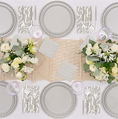 Overhead view of a table set for four with Sophistiplate’s Eco-Friendly Table Setting- Concrete: compostable scalloped plates, abstract-patterned napkins, silver cutlery, clear glasses, and white floral centerpieces on a woven beige runner.