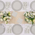 Overhead view of a table set for four with Sophistiplate’s Eco-Friendly Table Setting- Concrete: compostable scalloped plates, abstract-patterned napkins, silver cutlery, clear glasses, and white floral centerpieces on a woven beige runner.