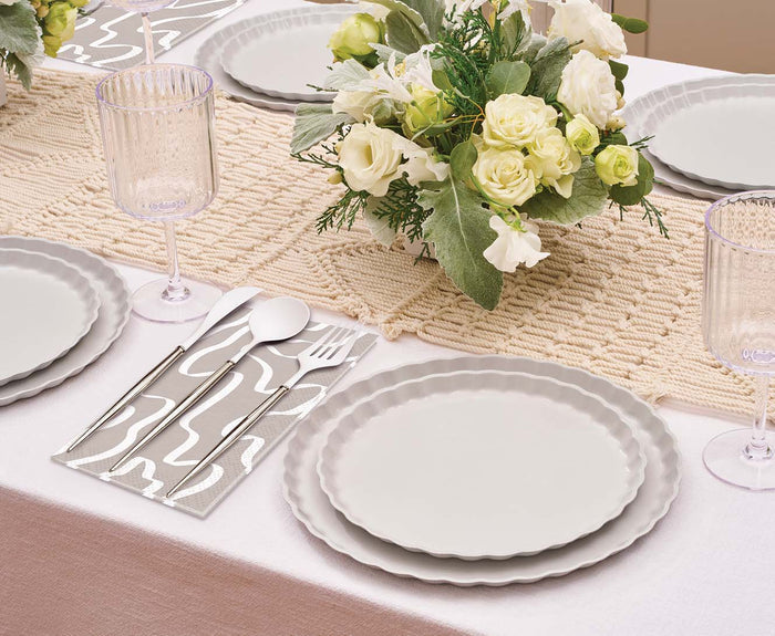A formal table is set with Sophistiplate's Eco-Friendly Table Setting- Concrete: stacked compostable plates, clear ribbed glasses, silver cutlery on a patterned napkin, and white roses with greenery on a woven runner.