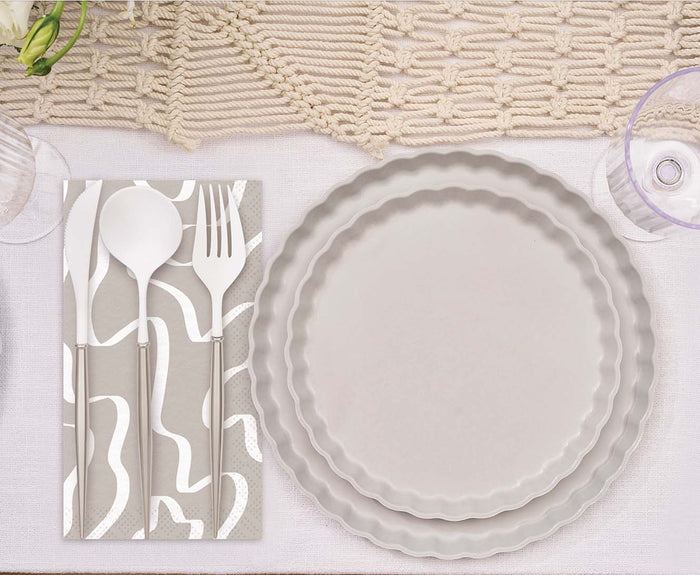The Eco-Friendly Table Setting-Concrete by Sophistiplate features two stacked beige compostable plates, a patterned napkin, white cutlery, and a clear ribbed glass on a woven runner. A white flower appears in the top left corner.