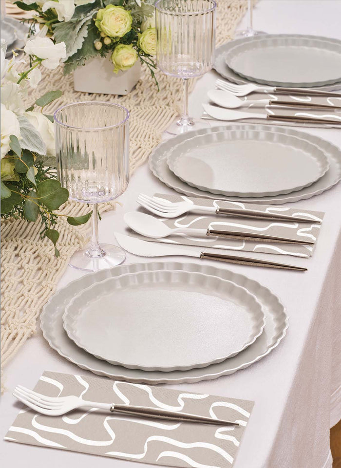 A close-up of Sophistiplate’s Eco-Friendly Table Setting- Concrete features gray plates, silverware on patterned napkins, ribbed glass goblets, and white floral centerpieces atop a woven runner.