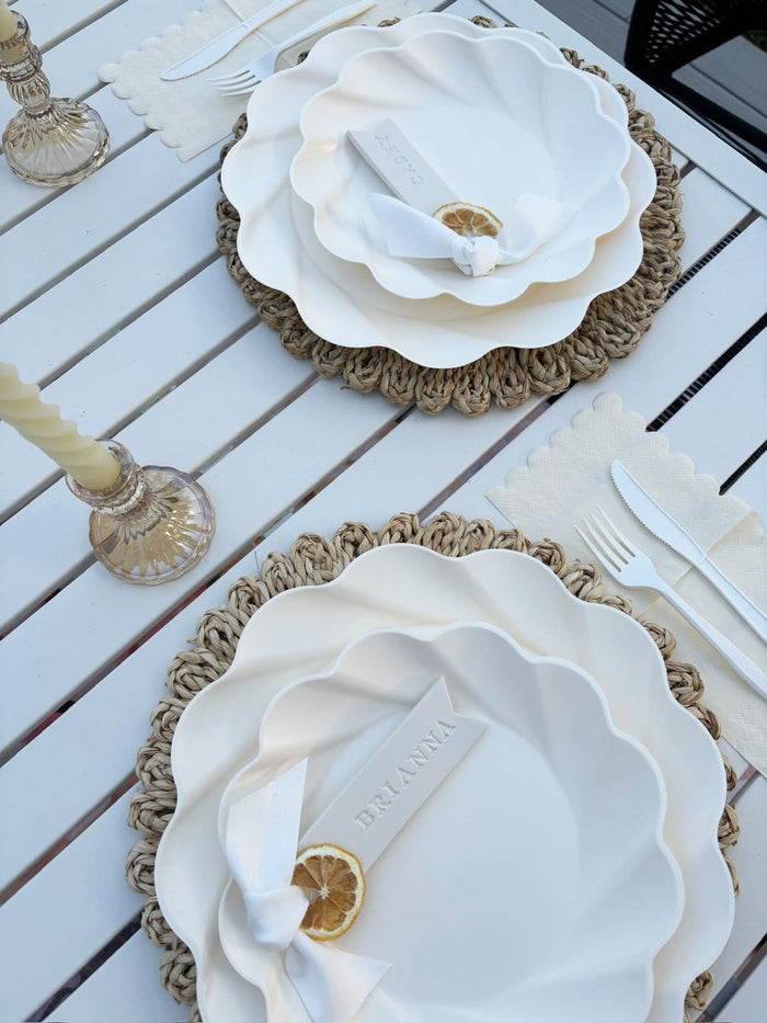 The Sophistiplate Eco-Friendly Table Setting- Cream includes a white outdoor table set with woven placemats, scalloped cream tableware, linen napkins, matching flatware, glass candle holders, and each setting features a lemon slice and ribbon-tied name card.