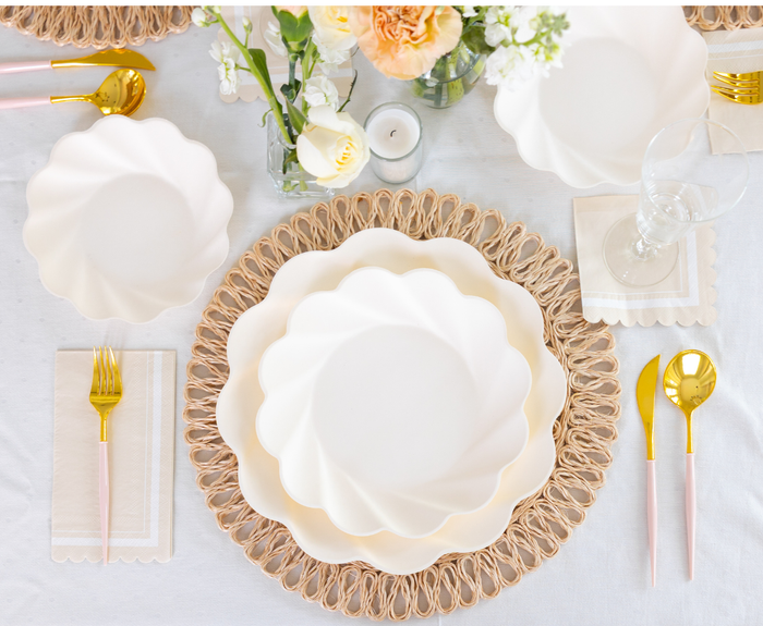 The Sophistiplate Eco-Friendly Table Setting- Cream features scalloped white compostable plates, gold and pink cutlery, napkins, a woven placemat, clear glassware, and floral centerpieces atop a white tablecloth.