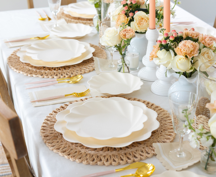 The Eco-Friendly Table Setting - Cream by Sophistiplate features white and gold scalloped compostable party plates, gold cutlery, woven placemats, pastel napkins, floral centerpieces in white vases, and tall pink candles for a chic tablescape.