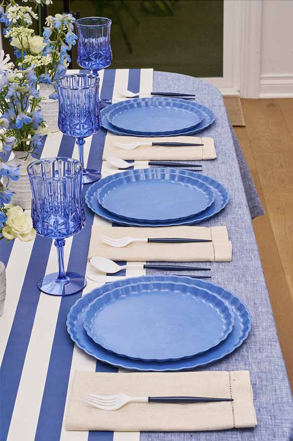 The Sophistiplate Eco-Friendly Table Setting—French Blue features compostable plates, blue glasses, cutlery in beige napkins, and a striped blue-and-white tablecloth, with blue and white flowers as a decorative touch.