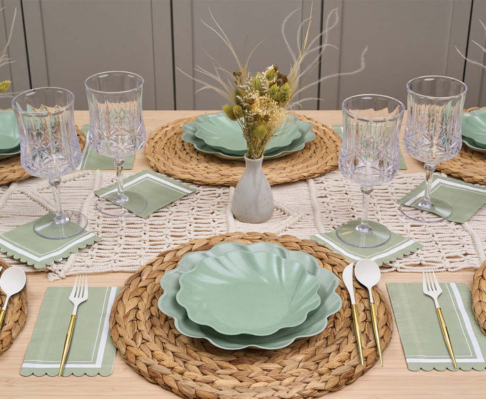 Set a beautiful table for four with the Eco-friendly Table Setting- Sage by Sophistiplate LLC, featuring green scallop plates, crystal glasses, gold cutlery on green napkins, woven placemats, and a dried flower vase centerpiece.