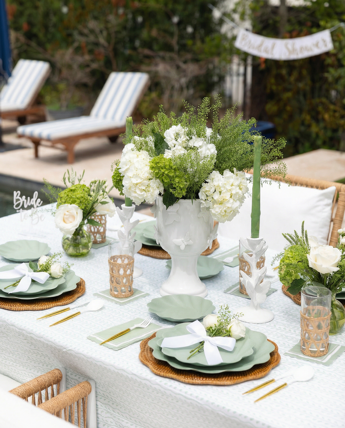 Create an elegant outdoor look with the Eco-friendly Table Setting- Sage by Sophistiplate LLC—featuring green and white accents, wicker placemats, green napkins, disposable plates, glasses, and a "Bridal Shower" banner on a white tablecloth.