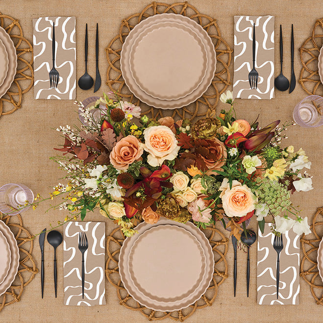 A top-view of the Sophistiplate Eco-Friendly Table Setting - Sand, featuring beige bamboo biodegradable plates, black cutlery, patterned napkins, clear glasses, and autumn-toned flowers on a burlap tablecloth.