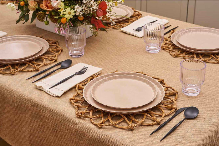 The Sophistiplate Eco-Friendly Table Setting- Sand features beige plates, black cutlery on white napkins, clear glasses, and a floral centerpiece in a white box atop a burlap tablecloth with woven placemats.
