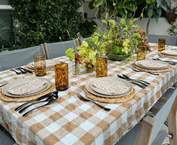 Create an inviting outdoor dining experience for six with Sophistiplate's Eco-Friendly Table Setting- Sand, featuring beige compostable plates, woven placemats, black cutlery, amber glasses, and a green and yellow floral centerpiece.