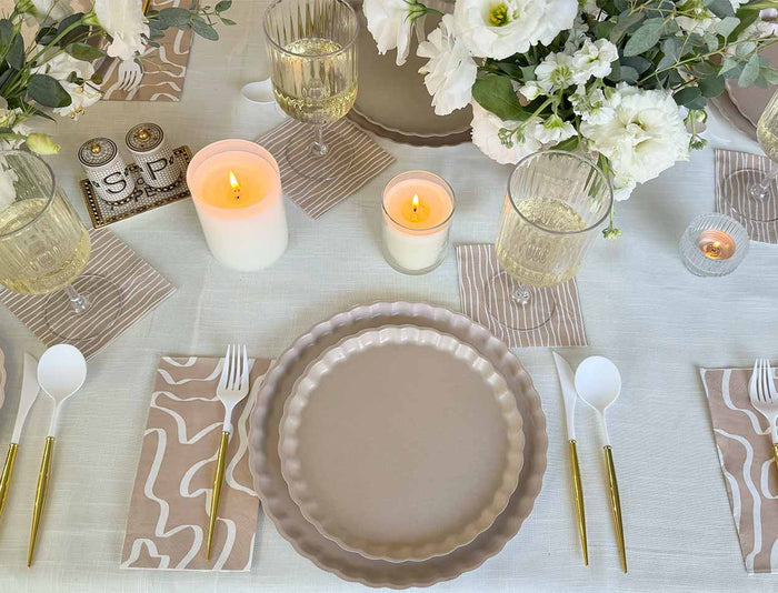 A table set for a meal with Sophistiplate’s Eco-Friendly Table Setting- Sand, featuring scalloped beige plates, gold and white cutlery, white wine glasses, candles, white florals, and abstract-patterned napkins and coasters on a white tablecloth.