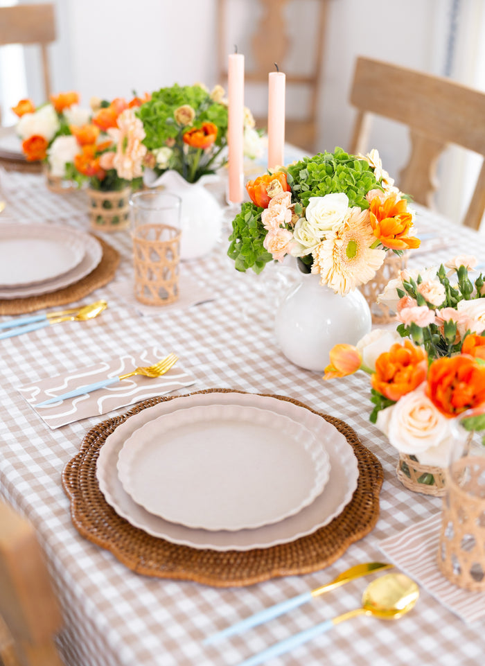 A beautifully set table featuring Sophistiplate’s Eco-Friendly Table Setting- Sand, with a checkered tablecloth, floral arrangements, pink candles, and biodegradable bamboo plates at every seat.