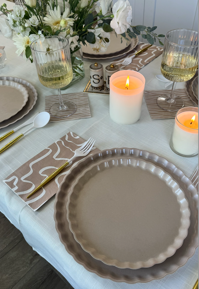 Create a stylish tablescape with the Sophistiplate Eco-Friendly Table Setting- Sand, featuring beige scalloped plates, gold cutlery, wine glasses, candles, neutral napkins, and a vase of white flowers.