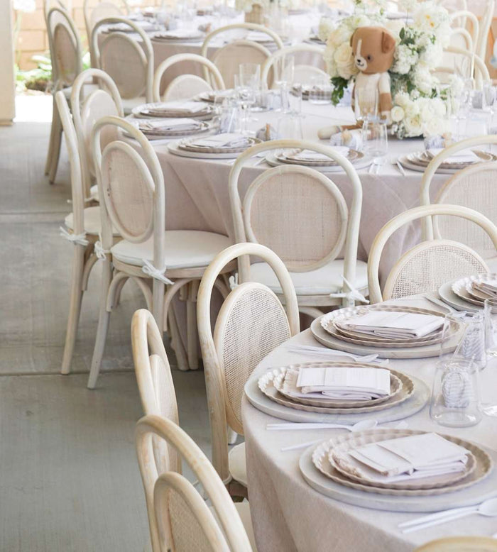 Elegant round tables set for an event with Sophistiplate's Eco-Friendly Table Setting- Sand, beige linens, white chairs, and a centerpiece of white flowers and a plush teddy bear.
