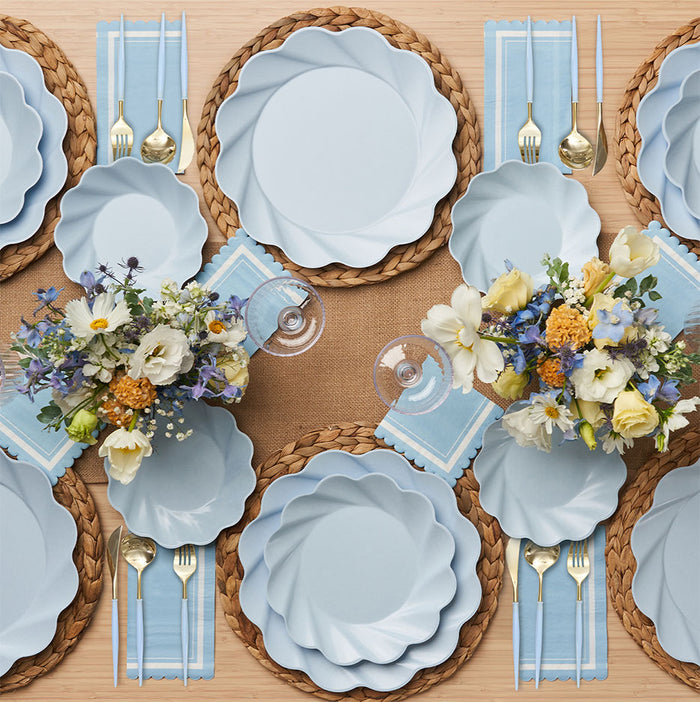 The Sophistiplate Eco-Friendly Table Setting- Sky Blue includes plates for four, gold cutlery, blue striped napkins, woven placemats, and two floral centerpieces with yellow, white, and purple blooms.