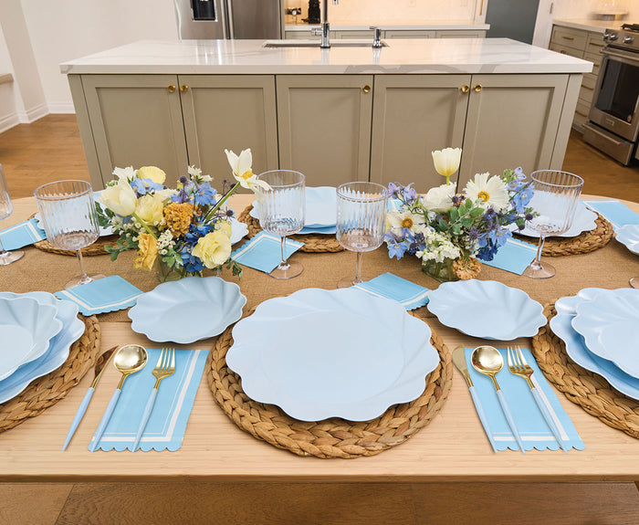 The Eco-Friendly Table Setting - Sky Blue by Sophistiplate features blue plates, woven placemats, gold flatware, blue napkins, and clear glasses. Yellow and white floral centerpieces add a fresh touch in a modern kitchen.
