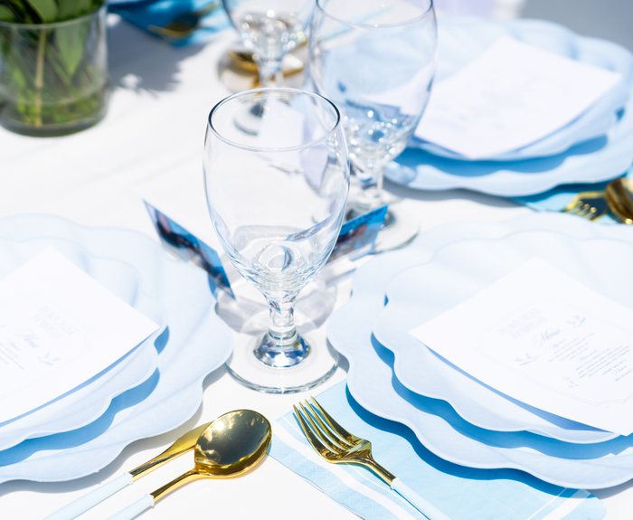 A close-up of a table set for a meal with Sophistiplate's Eco-Friendly Table Setting in Sky Blue, featuring clear wine glasses, gold and white cutlery, elegant plates with menus on top, all on a white tablecloth in bright sunlight.