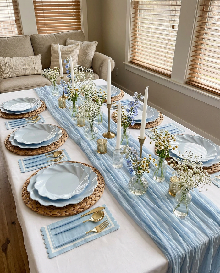 The Eco-Friendly Table Setting- Sky Blue by Sophistiplate features bamboo plates, gold cutlery, woven placemats, white candles, and vases with white and blue flowers on a low table for six. A beige sofa and window blinds are in the background.
