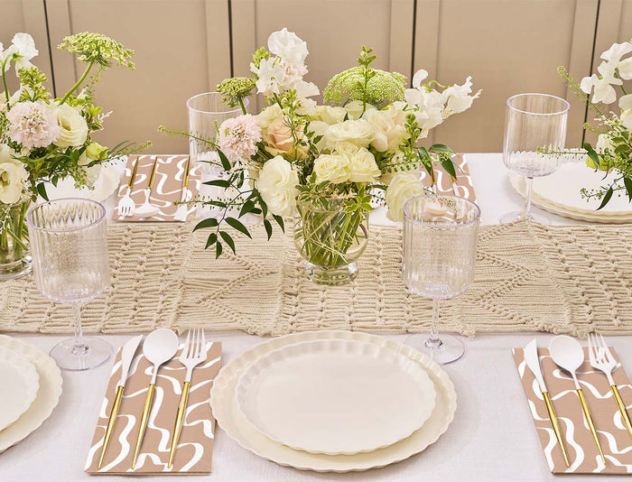 The Eco-Friendly Table Setting- White by Sophistiplate seats four with cream disposable plates, textured glassware, patterned napkins, and floral centerpieces on a beige woven runner—an elegant, neutral look with sustainable touches.