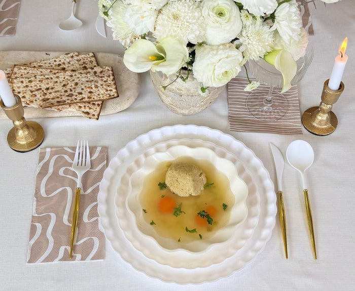 A bowl of matzo ball soup sits on a white tablecloth, surrounded by gold utensils, matzo, flowers, candles in gold holders, and Sophistiplate’s Eco-Friendly Table Setting- White for a refined and sustainable tablescape.