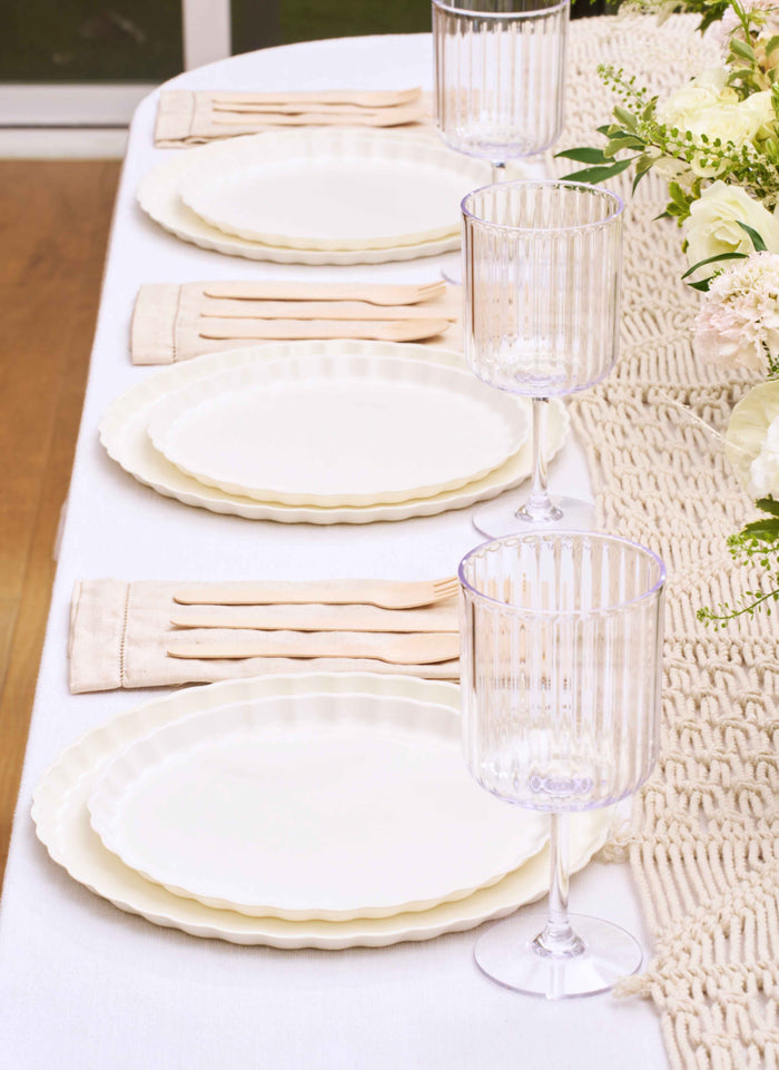 Create an elegant table setting with Sophistiplate’s Eco-Friendly Table Setting-White, featuring disposable plates, clear ribbed wine glasses, beige napkins with utensils, and a cream runner—accented by a white and green floral arrangement.