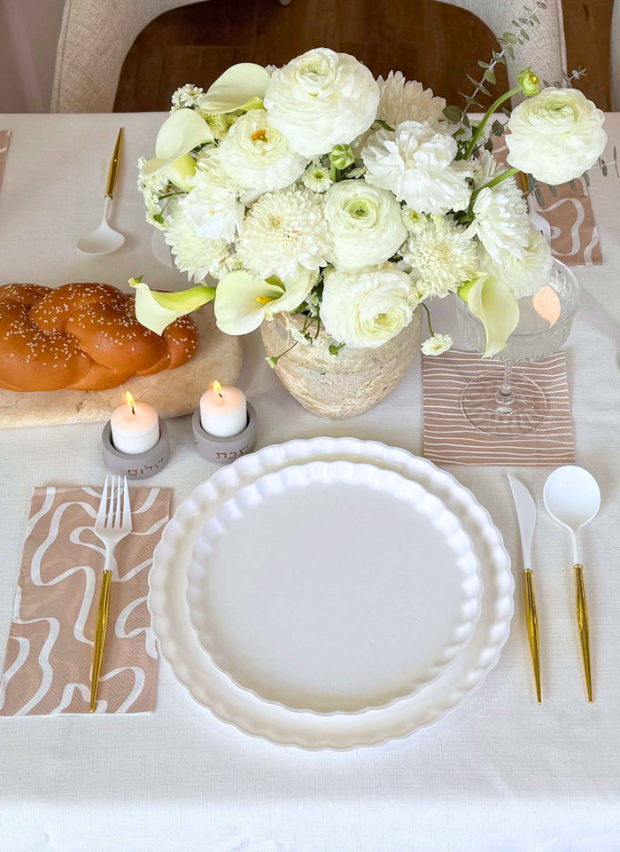 The Sophistiplate Eco-Friendly Table Setting-White features disposable plates, gold and white cutlery, white floral decor, candles, wine glasses, patterned napkins, and challah bread on a cream tablecloth.