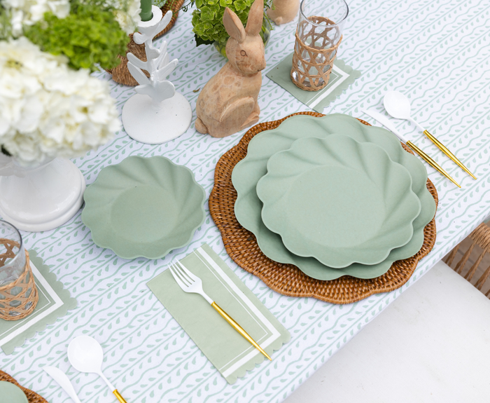 The Eco-friendly Table Setting- Sage by Sophistiplate LLC features disposable plates on woven chargers, gold and white cutlery, green napkins, wicker candle holders, white florals, wooden bunnies, and a patterned tablecloth.