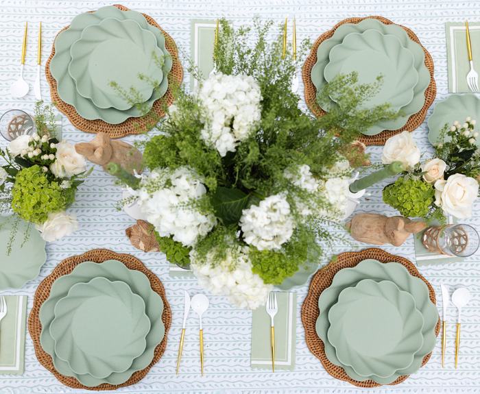 Create a spring tablescape with Sophistiplate LLC’s Eco-friendly Table Setting- Sage: green scalloped disposable plates, gold cutlery, woven placemats, white napkins, floral centerpieces, bunny accents, and a textured white tablecloth.