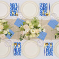 The Eco White & Blue Table Setting by Sophistiplate features eco-friendly cream plates, gold and blue cutlery, blue patterned napkins, wine glasses, and a centerpiece of white and pale pink flowers on a woven runner.