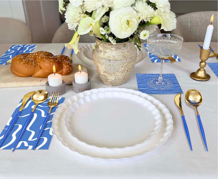 The Sophistiplate Eco White & Blue Table Setting features eco-friendly tableware, white plates, blue and white napkins, gold and blue cutlery, a vase of white flowers, candles, a wine glass, candlestick, and bread on a cutting board.